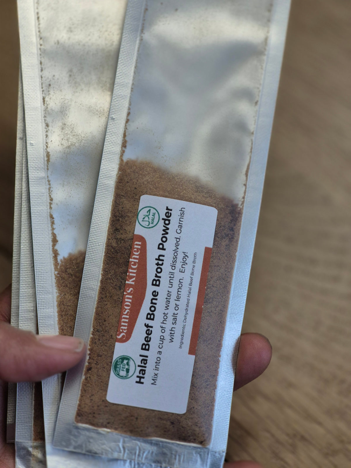 Packets of Samsons's Kitchen Halal Beef Bone Broth Powder held by a hand on a wooden surface.